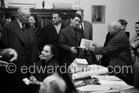 Pablo Picasso at a private viewing of his book illustrations in the Matarasso gallery in Nice, signing a book on his work, on the left standing (grey hair) Louis Aragon, sitting in the foreground Lilja Brik, sister of Elsa Triolet, behind with dark hat Nadine Léger, far right Jacqueline with Francine Weisweiller. Exposition "Pablo Picasso. Un Demi-Siècle de Livres Illustrés". Galerie H. Matarasso. 21.12.1956-31.1.1957. Nice 1956. - Photo by Edward Quinn