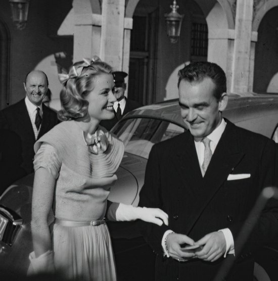 Prince Rainier and Princess Grace meeting Monegasque people in the courtyard of the Royal Palace for the official reception of the wedding presents. Louis Chiron in the background. Grace Kelly wore a pale blue chiffon day dress by the California designer James Galanos. She accessorized the dress with a small headpiece of matching blue ribbon, white gloves and the same Morabito needlepoint bag she had carried to the Academy Award of 1955. - Photo by Edward Quinn